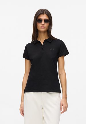 Kvinde iført sort poloshirt med korte ærmer og hvide bukser, med mørke solbriller, stående foran en ensfarvet lys baggrund.