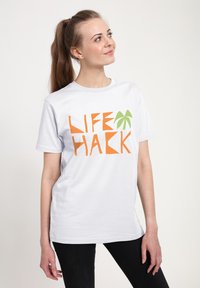 Hvid kortærmet t-shirt med orange og grøn "LIFE HACK" tekstdesign, med en palmeaccent, båret med sorte bukser.