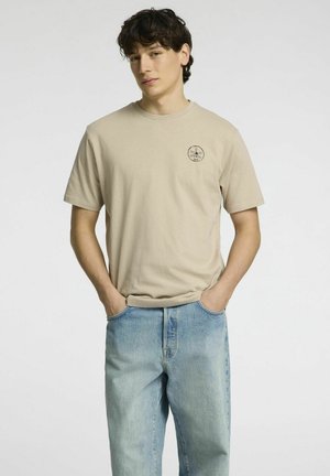 Beige bomulds t-shirt med korte ærmer og rund halsudskæring, med et lille sort grafisk motiv på brystet, parret med lyseblå denimjeans.