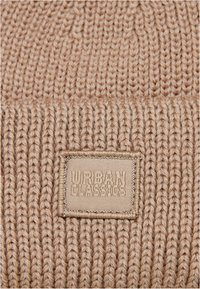 Világos bézs színű kötött sapka bordázott textúrával, elöl egy "URBAN CLASSICS" feliratos szövetcímkével.