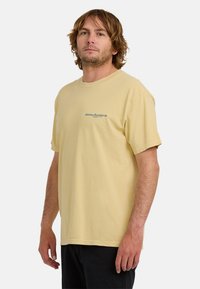 Żółta bawełniana koszulka z okrągłym dekoltem i krótkimi rękawami, z niebieskim nadrukiem na lewej części klatki piersiowej. Prosty, luźny krój.