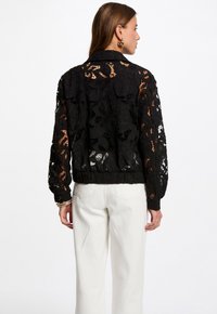 Veste en dentelle noire avec un motif floral, manches longues et taille cintrée. Portée sur un pantalon blanc, mettant en valeur le design complexe.