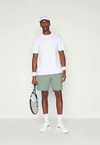 Ανδρική αθλητική εμφάνιση που περιλαμβάνει λευκό κοντομάνικο μπλουζάκι, πράσινα σορτς, λευκά περιβραχιόνια και ανοιχτό καπέλο, κρατώντας μια ρακέτα τένις.