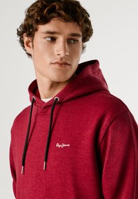 Tamsiai raudonas džemperis su gobtuvu, pagamintas iš minkštos medžiagos, su juostele su sidabriniais galiukais ir baltu logotipu kairėje krūtinės pusėje. Paprastas dizainas, be raštų.