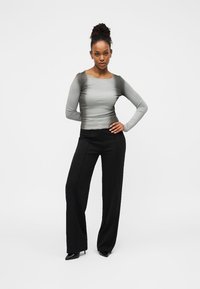 Γυναίκα με σκούρα μαλλιά που είναι δεμένα κότσο, φοράει εφαρμοστό γκρι μακρυμάνικο τοπ, μαύρο παντελόνι με φαρδιά πόδια και μαύρες μυτερές γόβες, στέκεται με το ένα χέρι στη μέση.