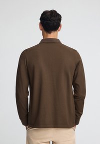 Brun långärmad polo skjorta med texturerad tyg, krage och sidslitsar, bärs med beige byxor, sedd bakifrån.