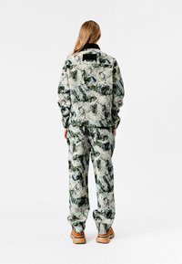 Chaqueta y pantalones con estampado floral en tonos verdes apagados, azules y beiges, hechos de una tela texturizada. Botas marrones completan el conjunto.