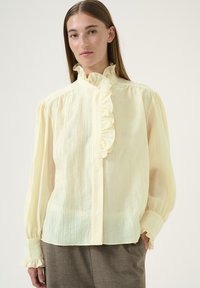 Ljusgul långärmad blus med en volangkrage, texturerad tyg och elastiska manschetter, i kombination med grå byxor.