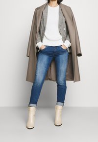 Abrigo beige extragrande, blazer gris a cuadros sobre suéter blanco de canalé, vaqueros azules y botines crema con hebillas.