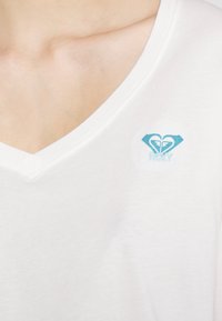 Valkoinen puuvillainen T-paita, jossa on V-kaula-aukko ja vasemmassa ylärinnassa pieni sininen Roxy-logo sydän- ja aaltoxenkuvioilla.