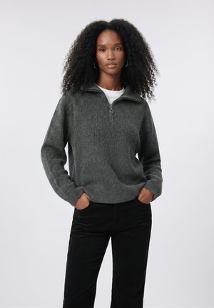 Femme aux cheveux bouclés portant un pull gris côtelé zippé et un pantalon noir, debout les mains dans les poches devant un fond uni.