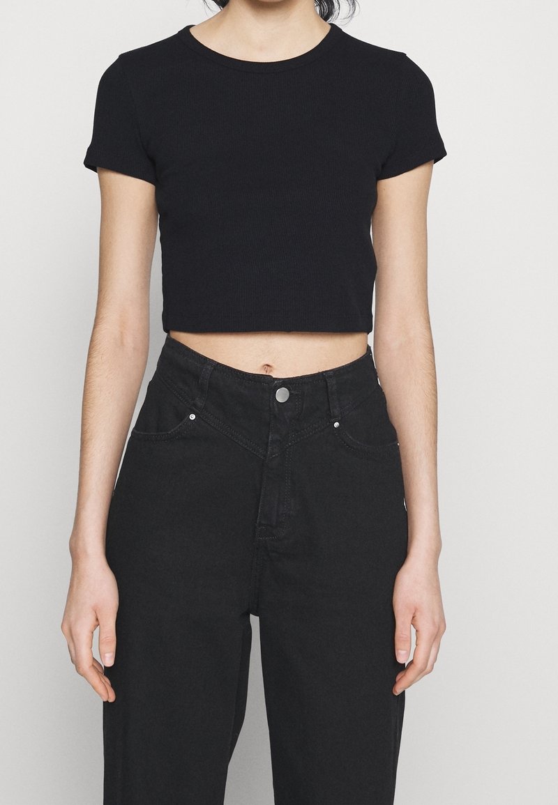 Personne portant un haut court noir à manches courtes et un jean noir taille haute, debout devant un fond blanc uni.