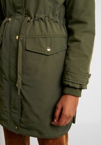 Khaki zelená parka se staženým pasem, kapsami na patentky, manžetovými rukávy a hladkým povrchem. Obsahuje zlaté kovové detaily.