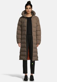 Frau steht mit einem langen braunen Daunenmantel über einem schwarzen Oberteil und Hosen sowie schwarzen Sneakers vor einem schlichten weißen Hintergrund.