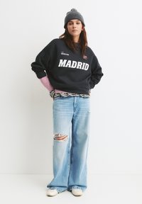 Čierna mikina s nápisom "MADRID" vytlačeným bielou farbou, skombinovaná so svetlomodrými roztrhnutými širokými džínsami. Doplnky tvoria šedá čiapka a ružové vrstvené manžety.