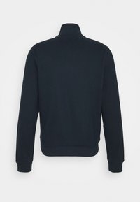 Laivansinisvetari, jossa on vetoketjukaulus, resorit hihansuissa ja helmassa. Liukas pinta ja minimalistinen muotoilu, ilman näkyviä logoja tai kuoseja.
