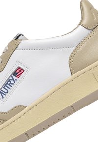 Sneaker in pelle bianca con accenti beige, suola in gomma texturizzata e patch brandizzato con una bandiera sul lato.