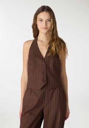 Giovane donna con lunghi capelli ondulati che indossa un gilet marrone a righe sottili senza maniche con bottoni e pantaloni abbinati a vita alta.