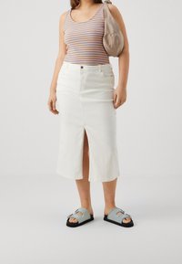 Falda de denim blanca con abertura frontal, combinada con una camiseta sin mangas de rayas multicolores. Los accesorios incluyen un bolso de hombro beige y sandalias azules.