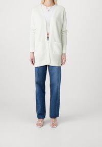 Cardigan long vert clair avec un motif texturé, ouverture devant, et deux poches avant, porté avec un jean bleu taille haute et des sandales claires.