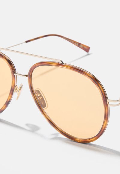 Primer plano de gafas de sol estilo aviador con lentes de tono ámbar y bordes de montura con patrón de carey sobre un fondo blanco.