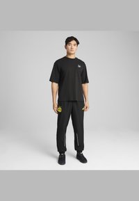 Camiseta negra de manga corta con un logo blanco, combinada con pantalones jogger negros que tienen un logo circular amarillo y acentos laterales en blanco. Zapatillas negras.