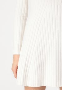Robe blanche à côtes avec des manches longues, présentant un tissu texturé, un ourlet évasé et des lignes verticales pour une dimension supplémentaire.