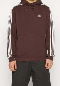 Sudadera marrón con rayas blancas a lo largo de las mangas, bolsillo canguro, capucha con cordón y pequeño logo blanco en el pecho. Material de algodón.