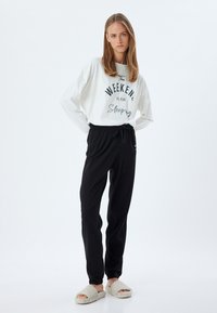 Top blanco de manga larga con estampado negro "El FIN DE SEMANA es para dormir", combinado con pantalones negros con cordón y zapatillas de casa cremosas y peludas.