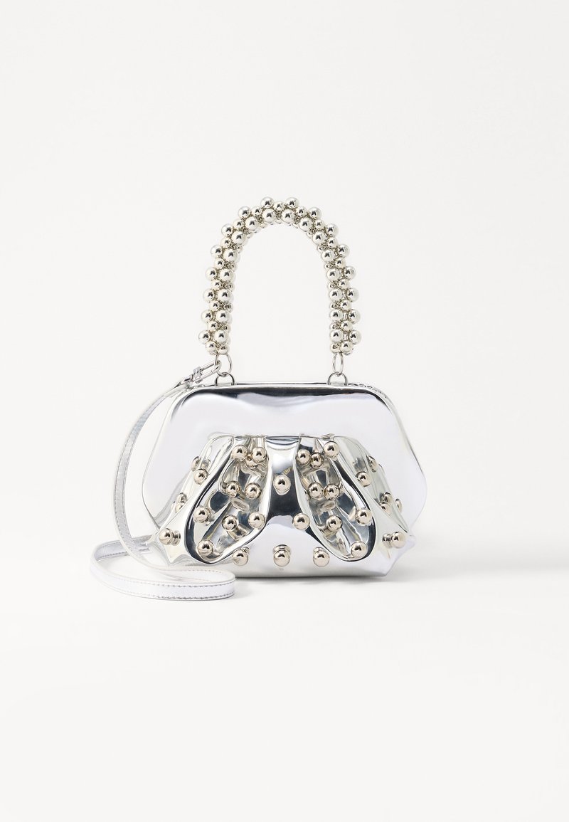 Strieborná metalická kabelka s okrúhlym tvarom, ozdobená guľovitými akcentmi a korálkovou rúčkou. Obsahuje odnímateľný ramenný popruh.