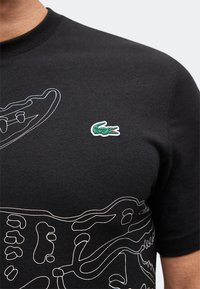 Μαύρο T-shirt με λευκό περίγραμμα σκελετού δεινοσαύρου και μικρό πράσινο λογότυπο κροκόδειλου στο στήθος.