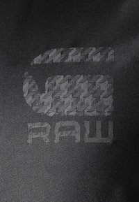 Svart tyg med en upphöjd textur och ett vävt mönster, där ordet "RAW" visas i ett fet, modernt typsnitt.