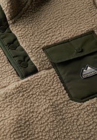 Beige fleece stof med struktureret overflade, prydet med olivengrønne accenter, knapper og en brystlomme med et logopatch.