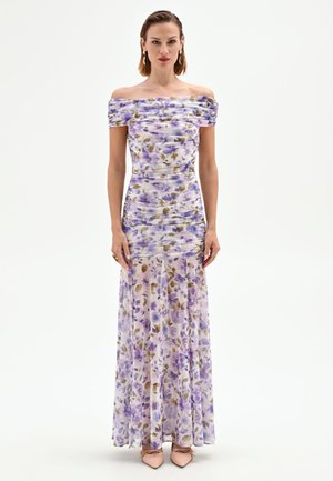 Femme debout portant une longue robe épaules dénudées avec un imprimé floral violet et marron et des chaussures pointues couleur beige sur fond blanc.