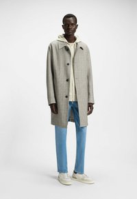 Cappotto a quadri chiari in tono beige e marrone, con colletto e tre bottoni, indossato sopra un hoodie beige, abbinato a jeans azzurri chiari e sneakers bianche.