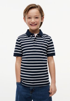 Usmievajúci sa chlapec so svetlohnedými vlasmi, oblečený v tmavomodro-biele pruhované polo tričko a modré rifle, stojaci pred jednofarebným pozadím.