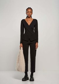 Top nero avvolgente con maniche lunghe, abbinato a pantaloni neri a vita alta. La modella tiene una borsa testurizzata beige chiaro e indossa scarpe nere massicce.