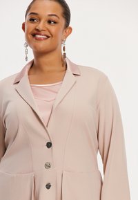 Lichtroze blazer met een enkele knoopsluiting, lange mouwen en een reverskraag, gedragen over een lichtroze top. Zilveren opvallende oorbellen.