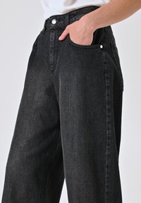 Zwarte denim broek met wijde pijpen en een hoge taille, voorzien van zijzakken en een knoopsluiting aan de voorkant. Textiel met een licht glanzende textuur.