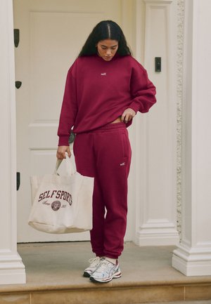 Woman in burgundy sweatshirt and sweatpants holding a white tote bag with "SELF SPORTS" text, standing on a doorstep in front of a white door.
