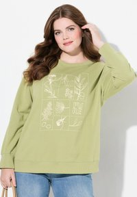 Felpa verde chiaro con un motivo a griglia di disegni botanici e la parola "PURE". Maniche lunghe e polsini a coste. Indossata con jeans blu.