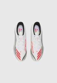 White athletic shoes with red accents, featuring a sleek design, laces, and black inner lining. Includes diagonal stripes and branding.