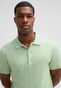 Jasnozielona koszulka polo z krótkim rękawem, wykonana z miękkiej dzianiny, z klasycznym kołnierzykiem i zapięciem na dwa guziki. Gładka tekstura, prosty design.