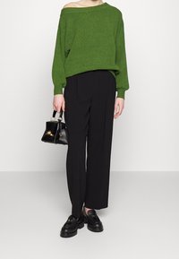 Groene oversized sweater met off-shoulder ontwerp, gecombineerd met zwarte wijde broek. In één hand wordt een kleine zwarte handtas vastgehouden.
