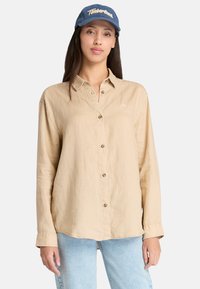 Camicia beige a maniche lunghe con bottoni, realizzata in tessuto leggero, caratterizzata da un sottile logo, con una vestibilità rilassata e orlo arrotondato. Indossata con jeans azzurri chiari.