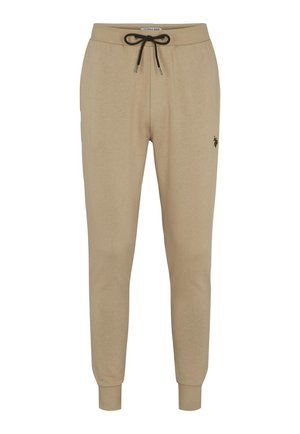 Homme portant un sweat-shirt beige à manches longues et un pantalon de jogging assorti avec des détails de logo, accompagné de baskets blanches, debout les mains détendues.