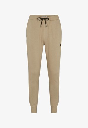 Homme portant un sweat-shirt beige à manches longues et un pantalon de jogging assorti avec des détails de logo, accompagné de baskets blanches, debout les mains détendues.