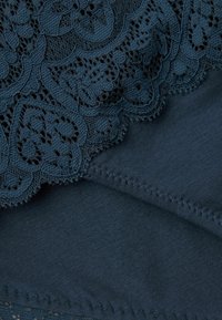 Tejido de color verde azulado oscuro con intrincado encaje en la parte superior que presenta patrones florales, y una textura suave y lisa debajo. Los bordes tienen costuras bien hechas.