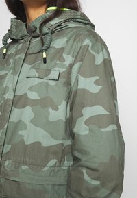 Camo jacket in shades of green with a hood, featuring drawstring accents and a chest pocket. Made from lightweight fabric with a smooth texture.