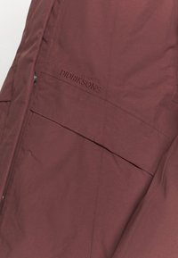 Gros plan sur la poche d'une veste bordeaux avec rabat, coutures surpiquées et le nom de la marque "Didriksons" brodé au-dessus de la poche.